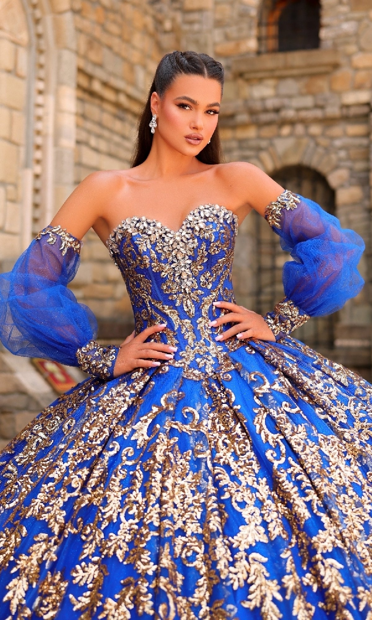 A woman poses in the Sequin Quinceanera Dress: Amarra 54238, a blue gown with gold embroidery, sheer puff sleeves, and a corset bodice, standing before a stone building.